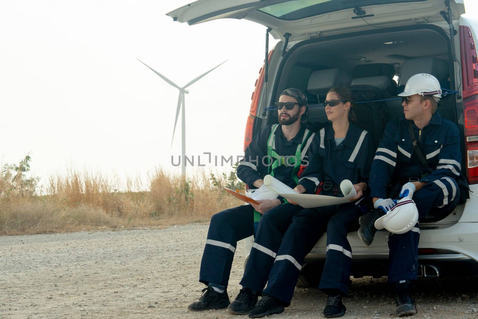 Close up group of technician workers sit on back of van and discuss about work in plan paper on the road with windmill or wind turbine on the background. by nrradmin