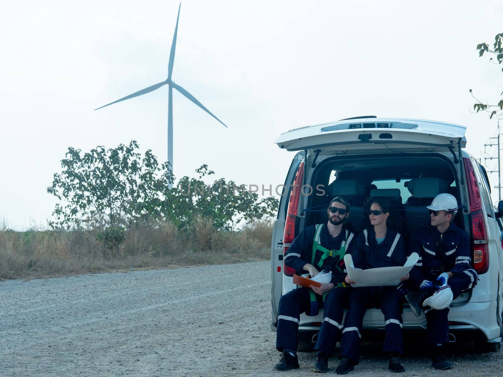 Group of technician workers sit on back of van and discuss about work in plan paper on the road with windmill or wind turbine on the background.