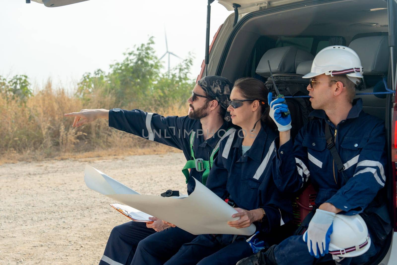 Group of technician workers sit on back of van and discuss about work in plan paper and one point to left side stay on the road in area of windmill or wind turbine on the background.