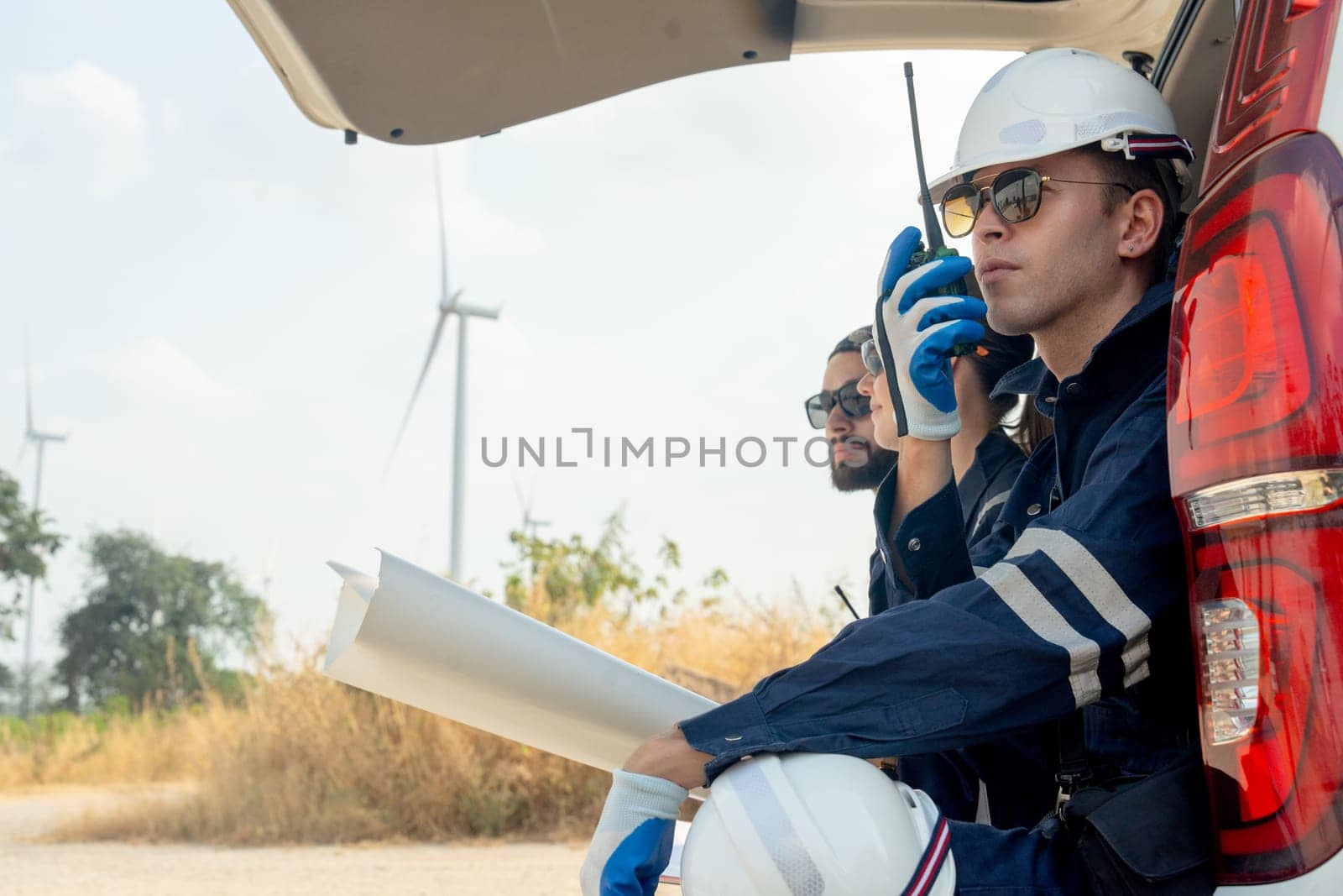 Close up technician worker man use walkie talkie to contact his co-worker and sit in the back of van in area of power plant with wind tubine or windmill.