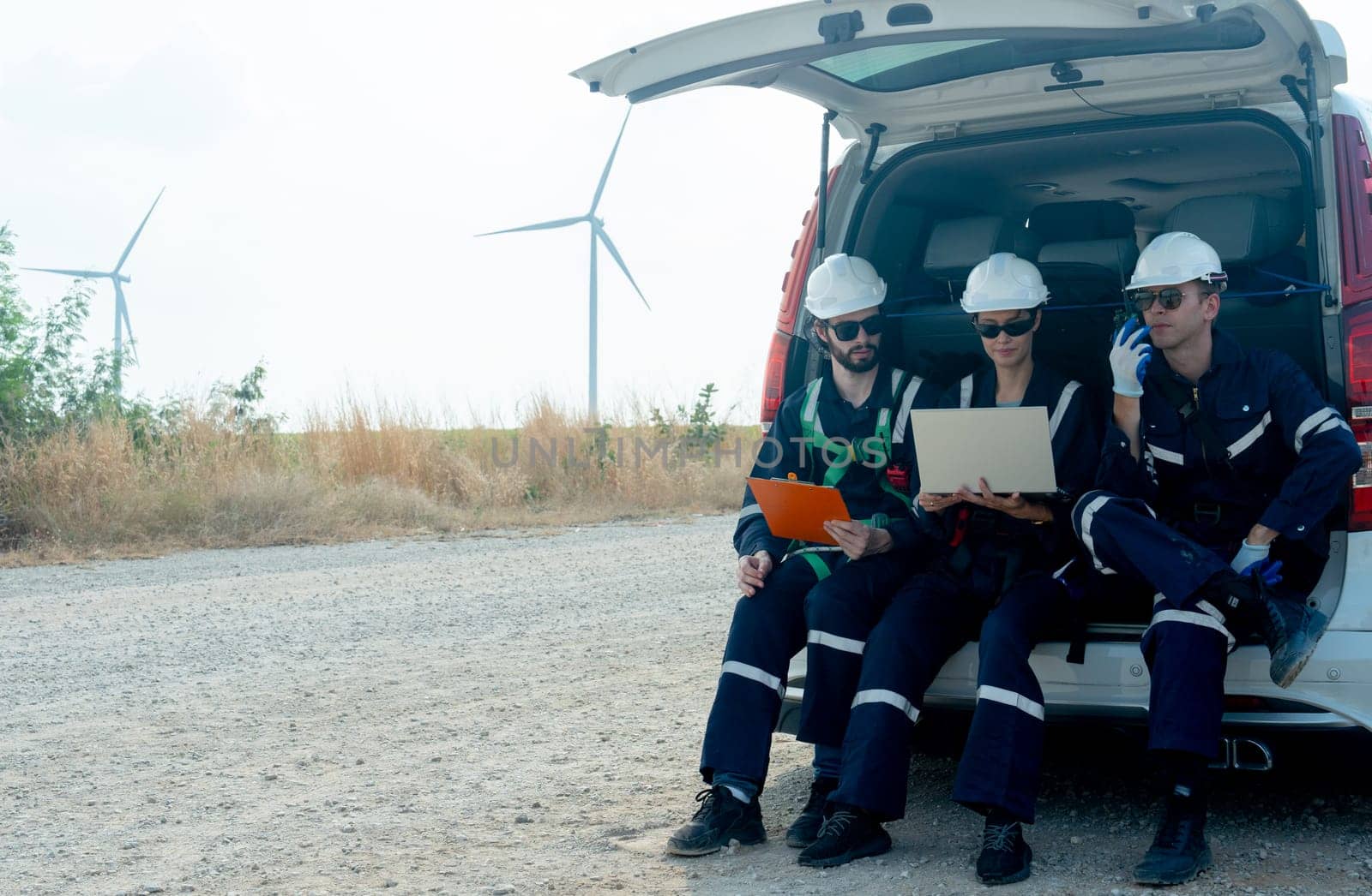 Group of technician workers sit on back of van on the road and discuss about work using laptop with windmill or wind turbine on the background. by nrradmin