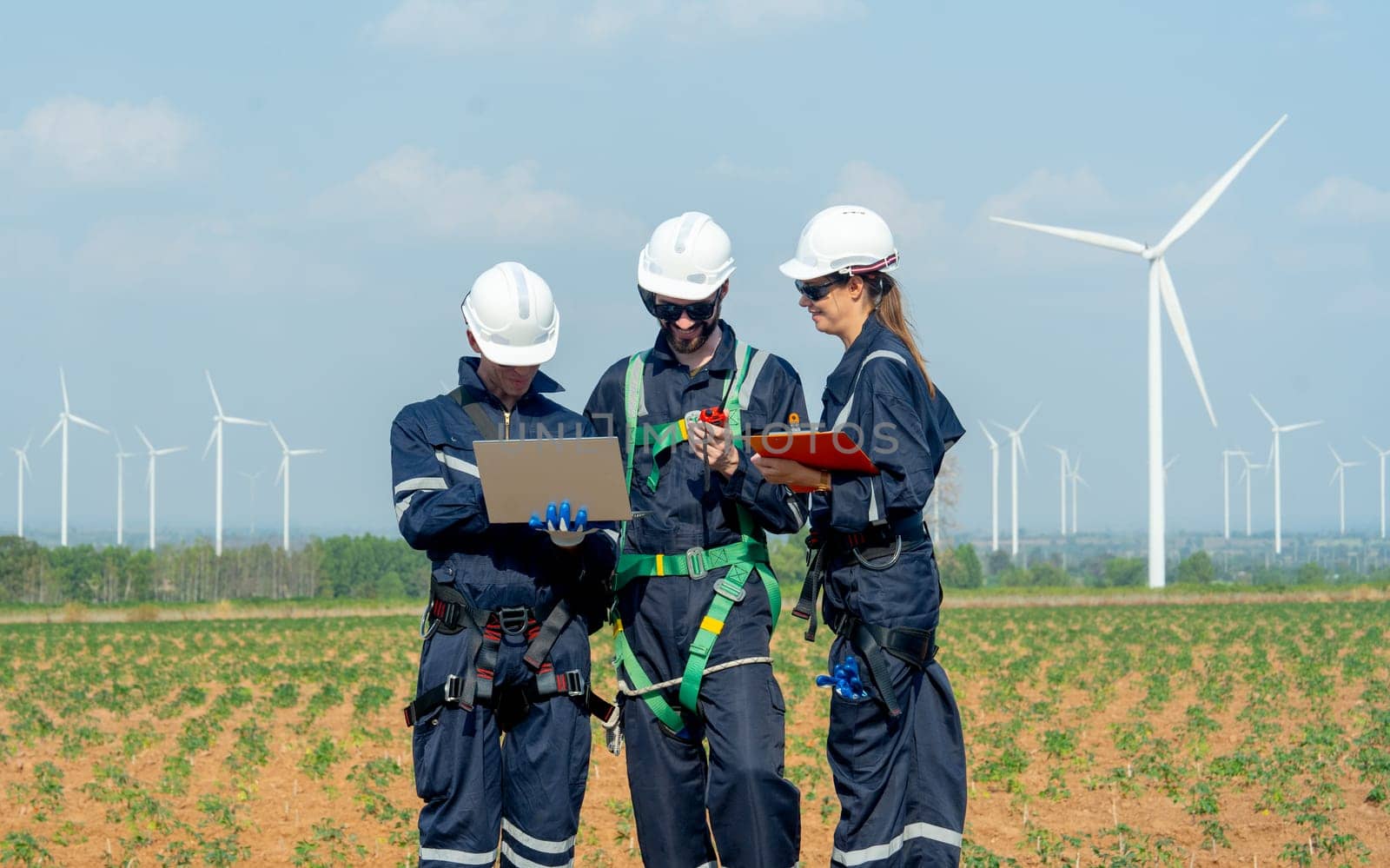 Close up professional technician workers man and woman stand in front of row of windmill or wind turbine and work together using laptop with day light and they look happy with smiling.