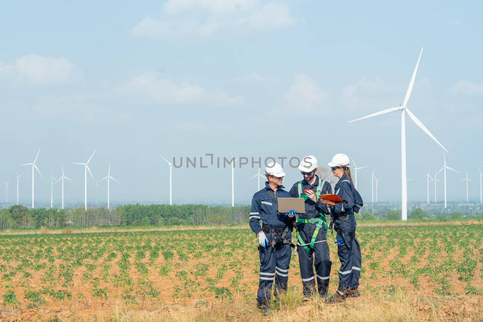 Side view technician workers man and woman stand in front of row of windmill or wind turbine and work together using laptop with day light.