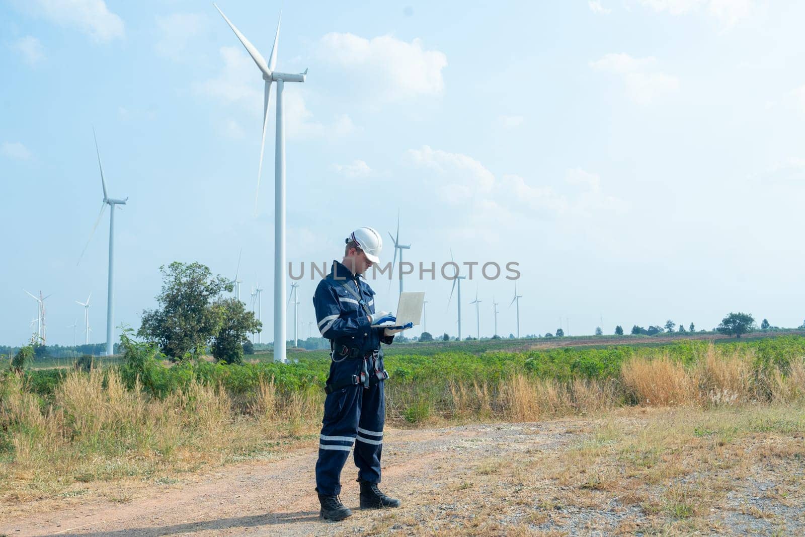 Wide shot of technician worker hold laptop and look to the monitor for working in front of windmill or wind turbine with blue skay in area of power plant factory.