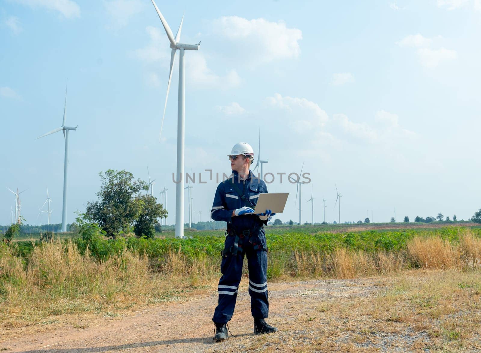 Wide shot of professional technician worker hold laptop and look to left side in front of windmill or wind turbine with blue skay in area of power plant factory.