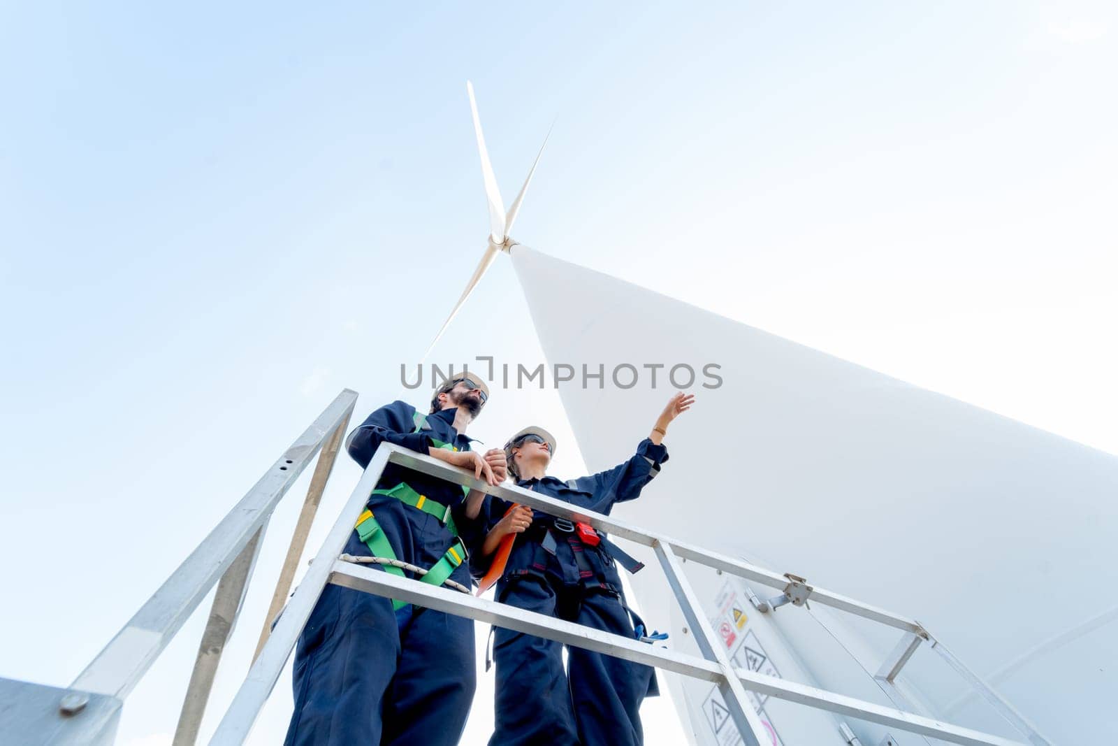 Lower view of professional technician man and woman stay on base of windmill or wind turbine and woman also point to the right with blue sky. by nrradmin
