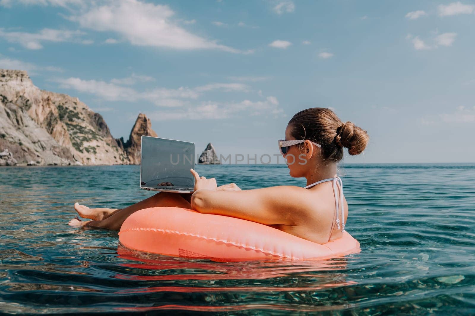 Woman freelancer works on laptop swimming in sea on pink inflatable ring. Pretty lady typing on computer while floating in the sea on inflatable donut at sunset. Freelance, remote work on vacation