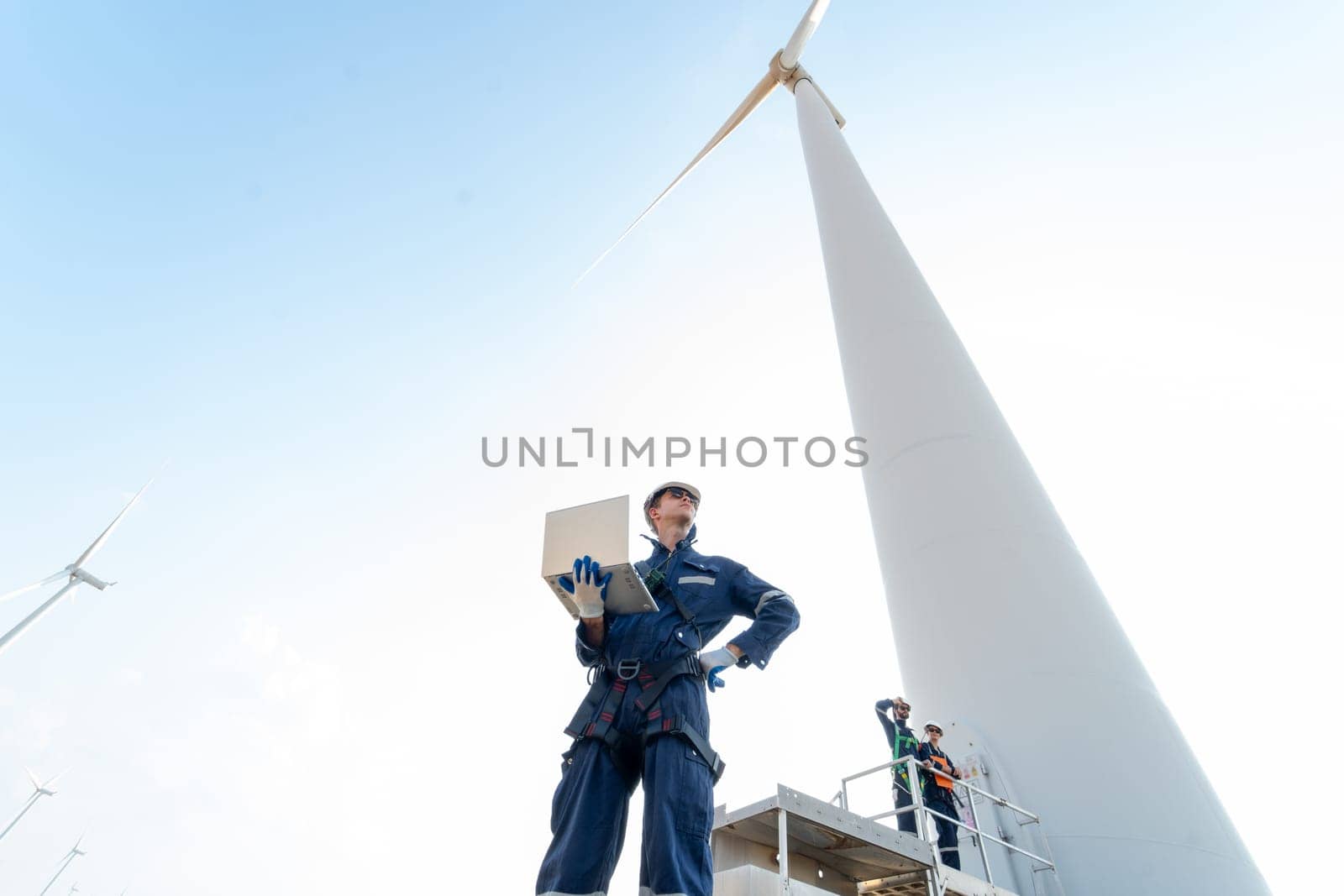 Wide shot professional technician man stand with hold laptop and stand near base of windmill or wind turbine also look to right side with his co-worker in the back. by nrradmin