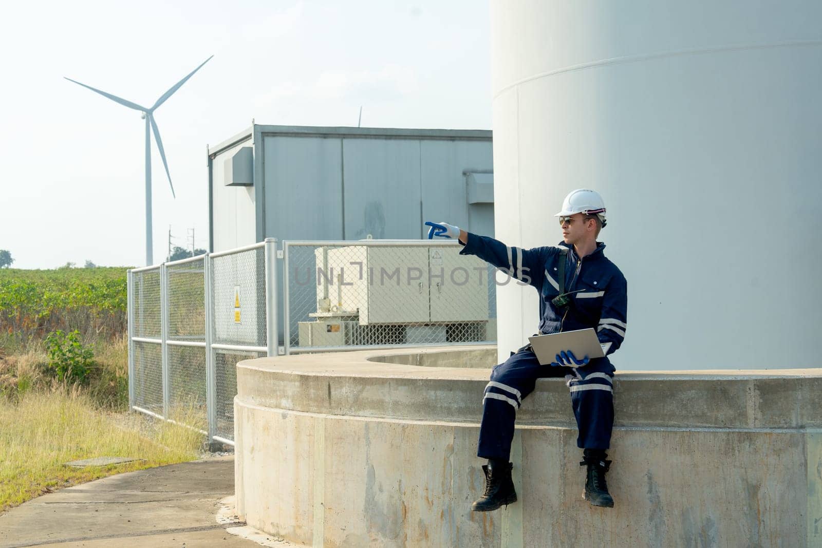 Professional technician worker hold laptop and sit on base of windmill or wind turbine also point to left side in area of green power plant.