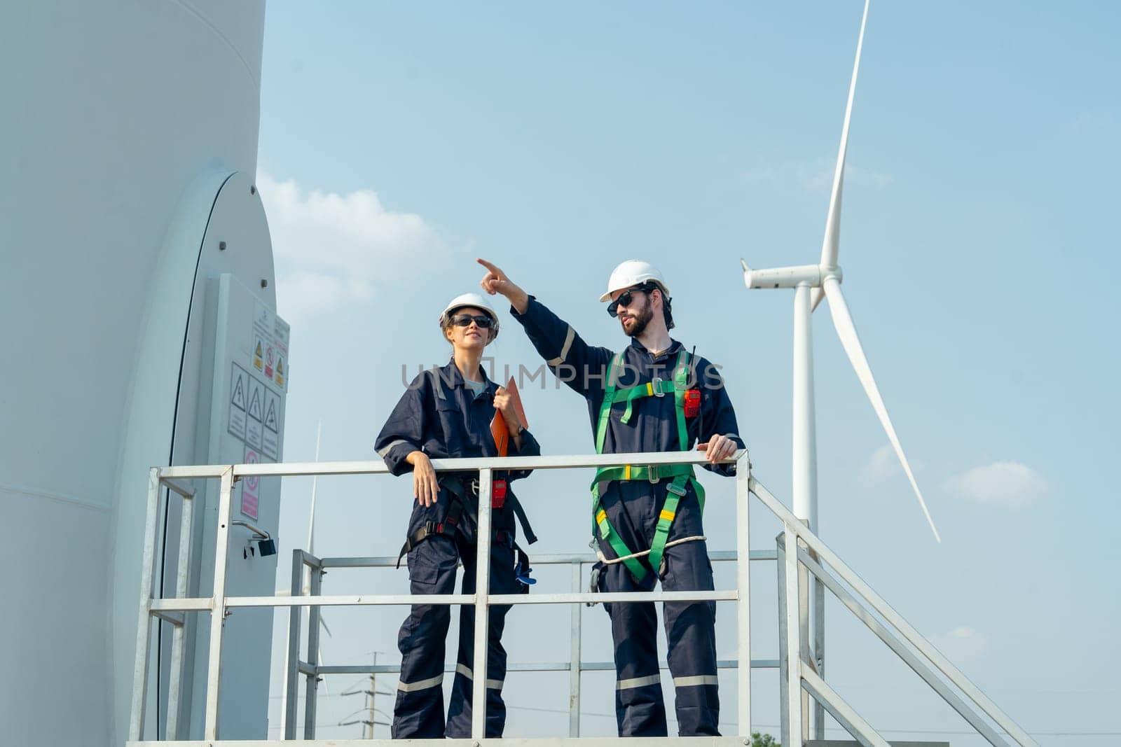 Two of professional workers stand and discuss about work with man also point to left side and stay in base of windmill or wind turbine in area of green power plant.
