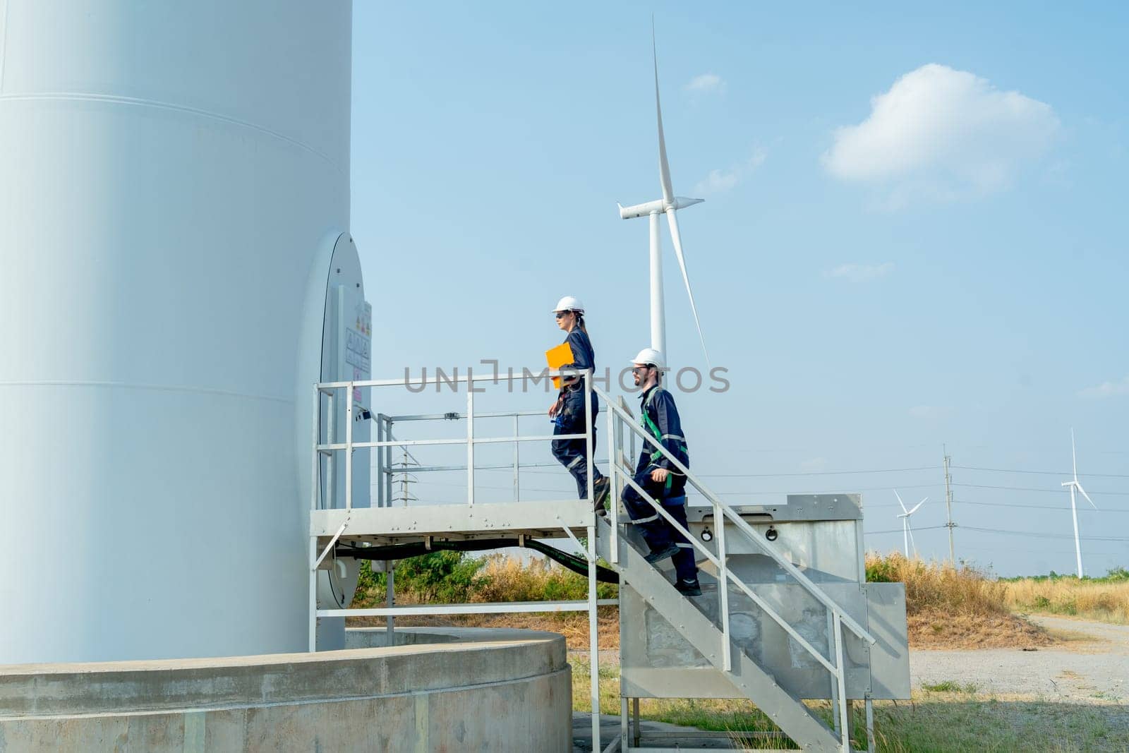 Two of technician workers walk up on stair of base of wind turbine or windmill to check or maintenance work in area of power plant.