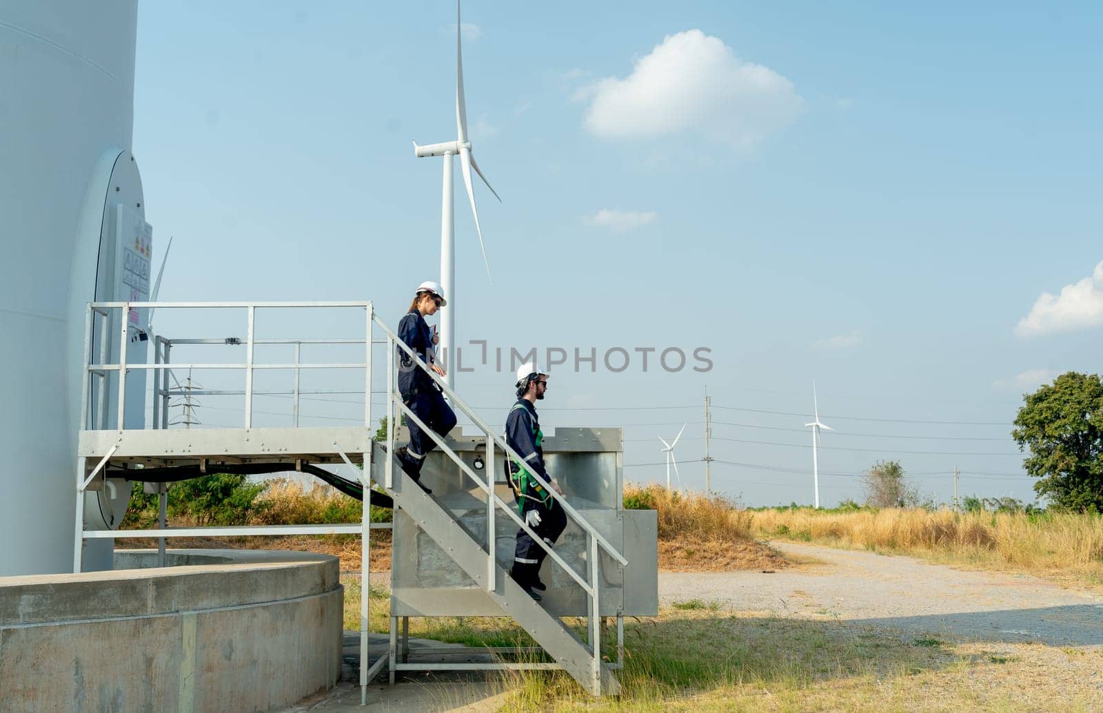 Two of technician workers walk down from stair of base of wind turbine or windmill after finish maintenance work in area of power plant.