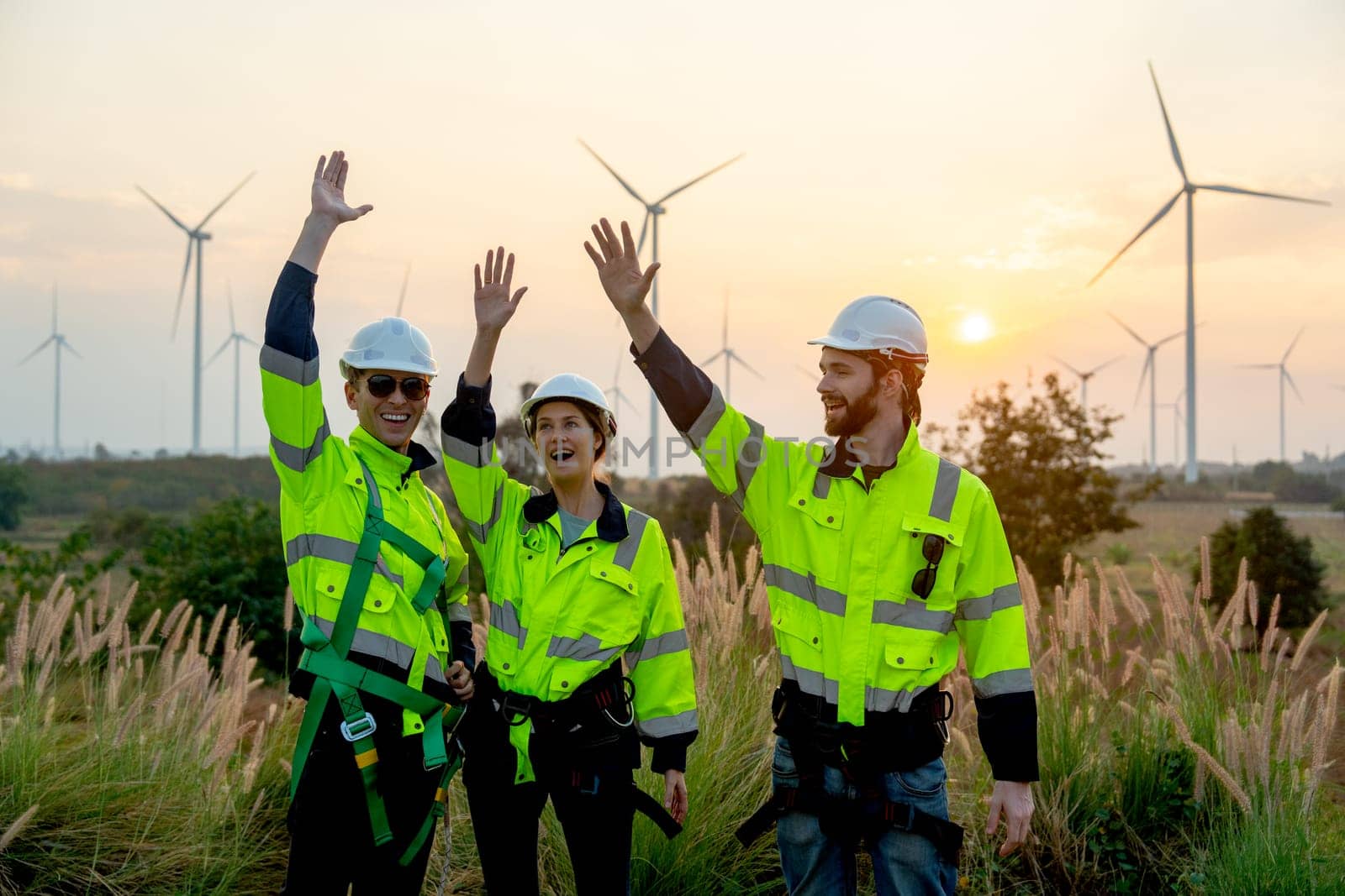 Group of technician worker man and woman stand with rest hands up express happiness during work and stand in front of windmill or wind turbine farm in area of power plant. by nrradmin