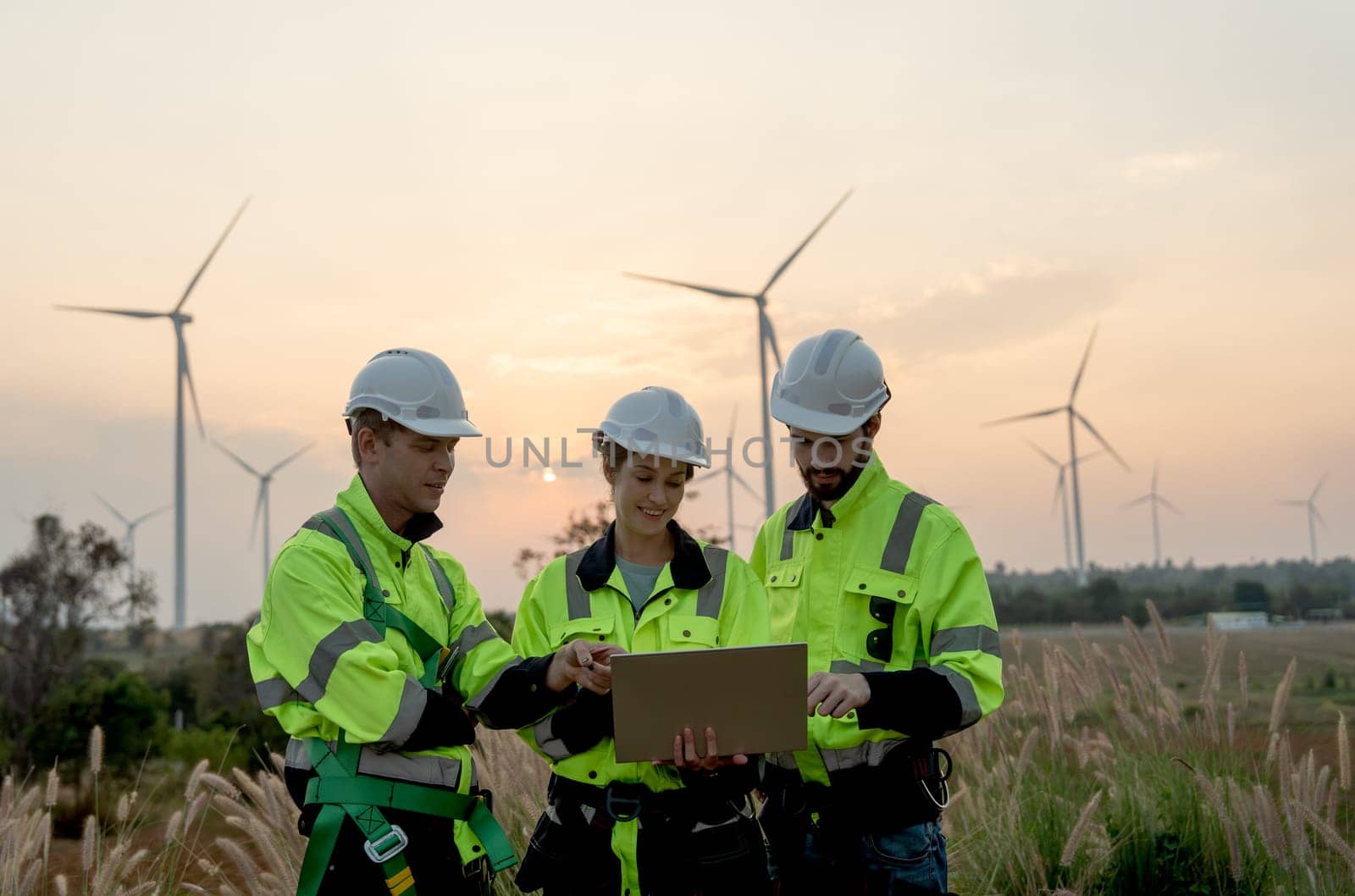 Group of professional technician workers man and woman work using laptop and stay in front of windmill or wind turbine with evening light. by nrradmin