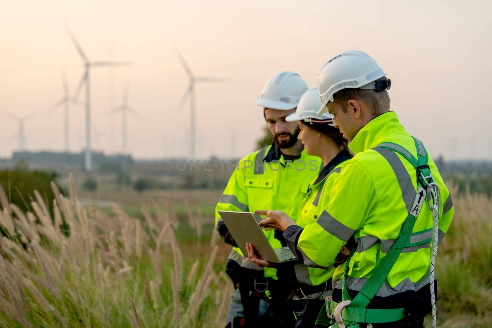 Side view of technician workers man and woman work using laptop and stay in front of windmill or wind turbine with evening light.