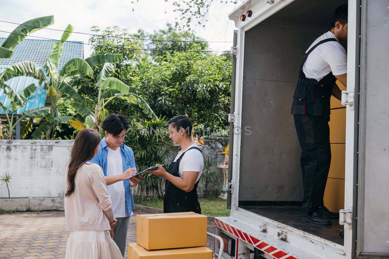 Professional movers assist a satisfied couple in signing the delivery checklist after furniture handling. Uniformed employees prioritize customer satisfaction. Moving Day Concept by Sorapop