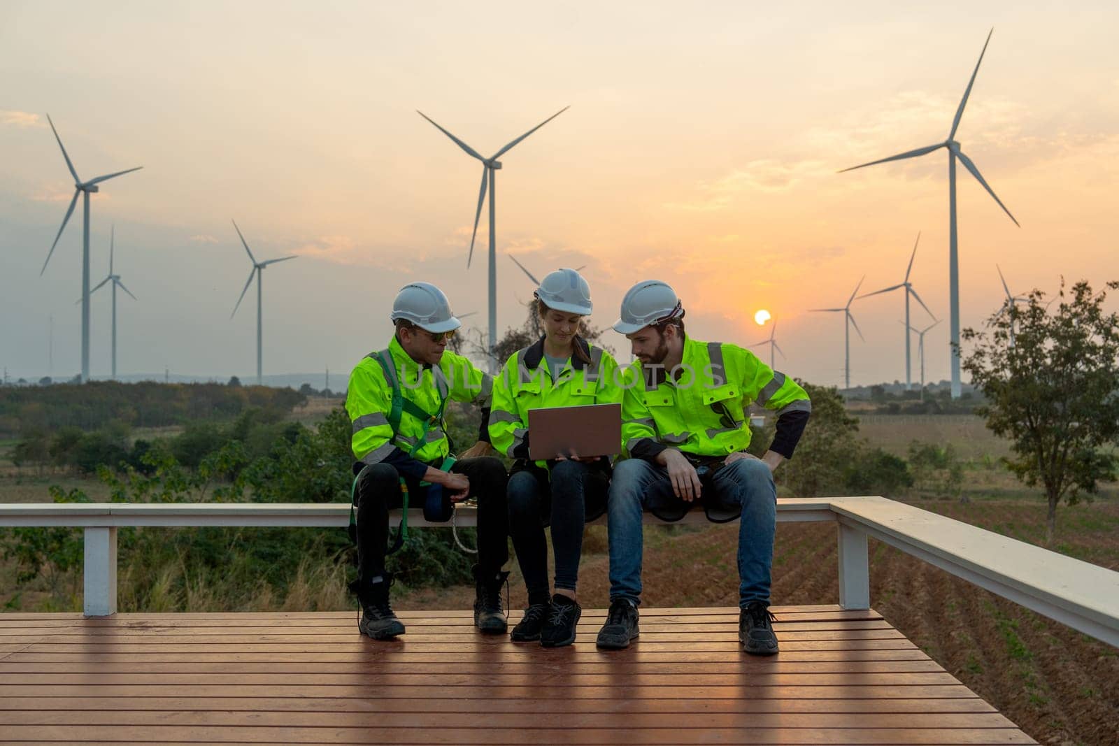 Group of professional technician workers with safety suit sit and discuss work using laptop and row of windmill or wind turbine on the background and evening light. by nrradmin