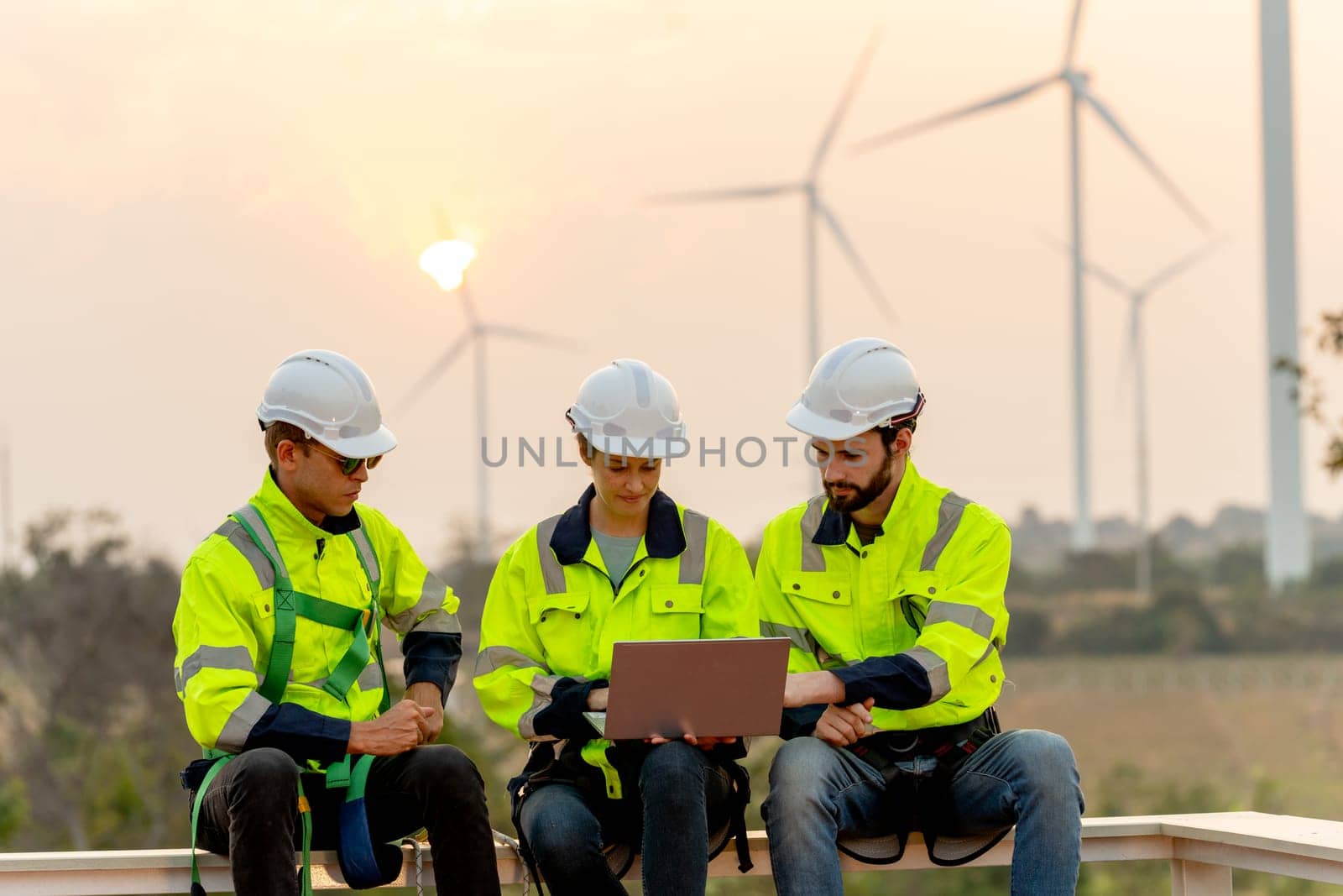 Close up group of professional technician workers with safety suit sit and discuss work using laptop and row of windmill or wind turbine on the background in evening.