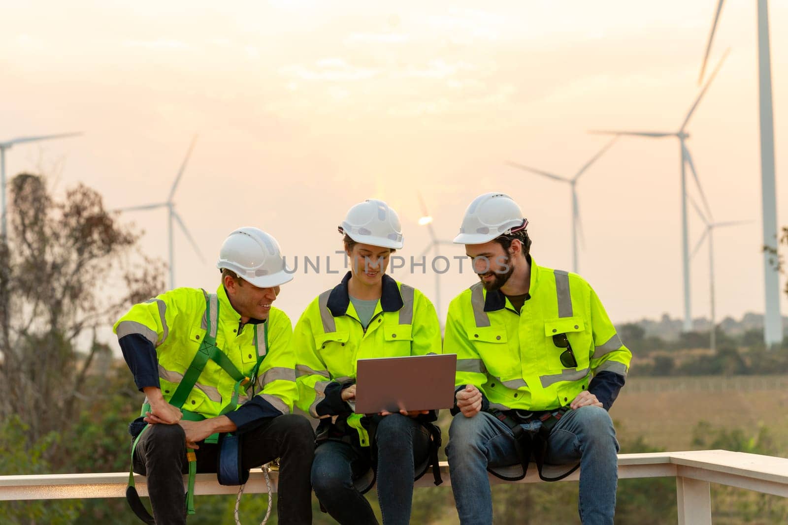 Group of professional technician workers with safety suit sit and discuss work using laptop and row of windmill or wind turbine on the background in evening. by nrradmin