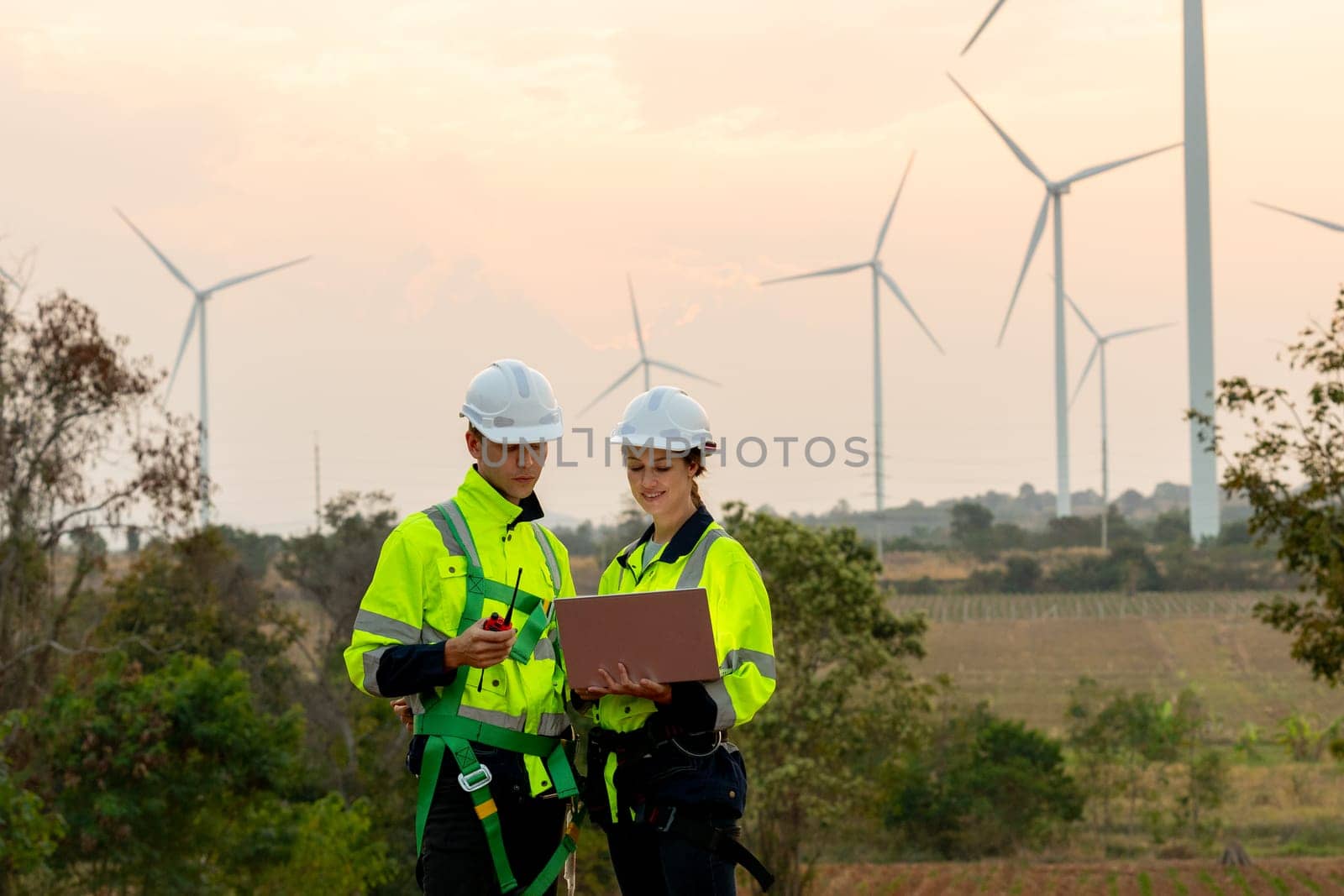 Professional technician worker man and woman with safety suit stand together using laptop to discuss work in front of windmill or wind turbine with evening light in area of power plant. by nrradmin