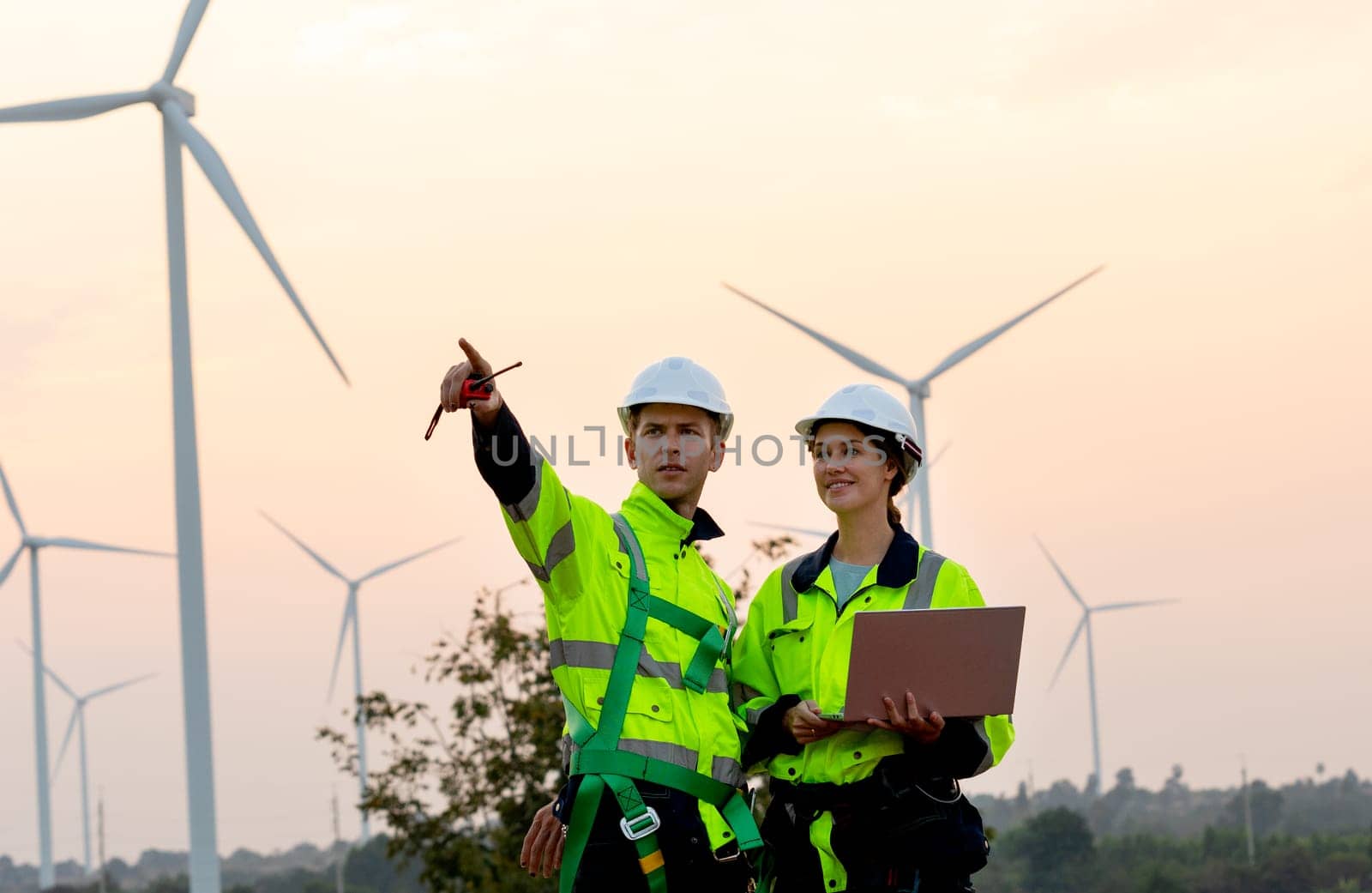 Technician workers use laptop to work and stay with her co-worker point to left side in evening with windmill or wind turbine on the background.