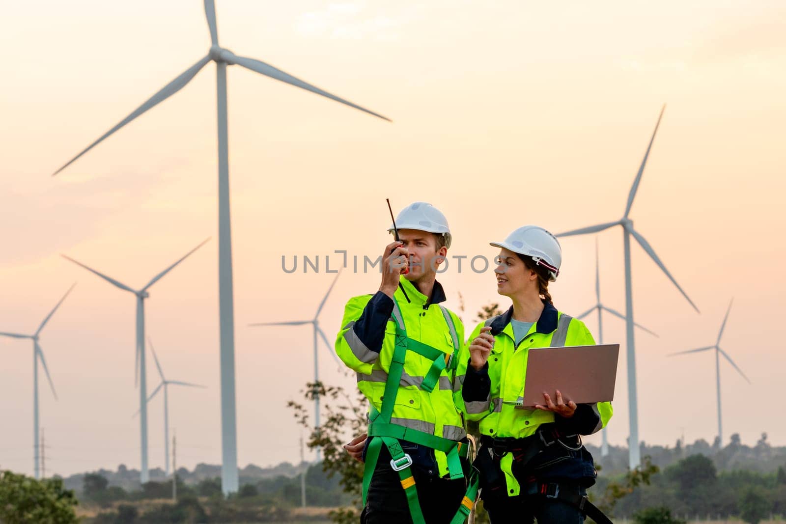 Professional technician man use walkie talkie to contact his co-worker and woman hold the laptop stand beside and work in front of windmill or wind turbine farm. by nrradmin