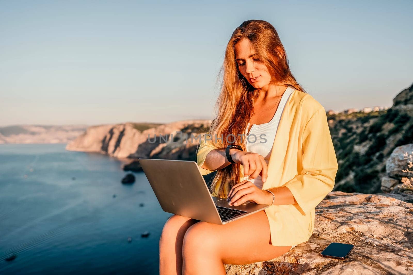 Digital nomad, Business woman working on laptop by the sea. Pretty lady typing on computer by the sea at sunset, makes a business transaction online from a distance. Freelance, remote work on vacation by panophotograph