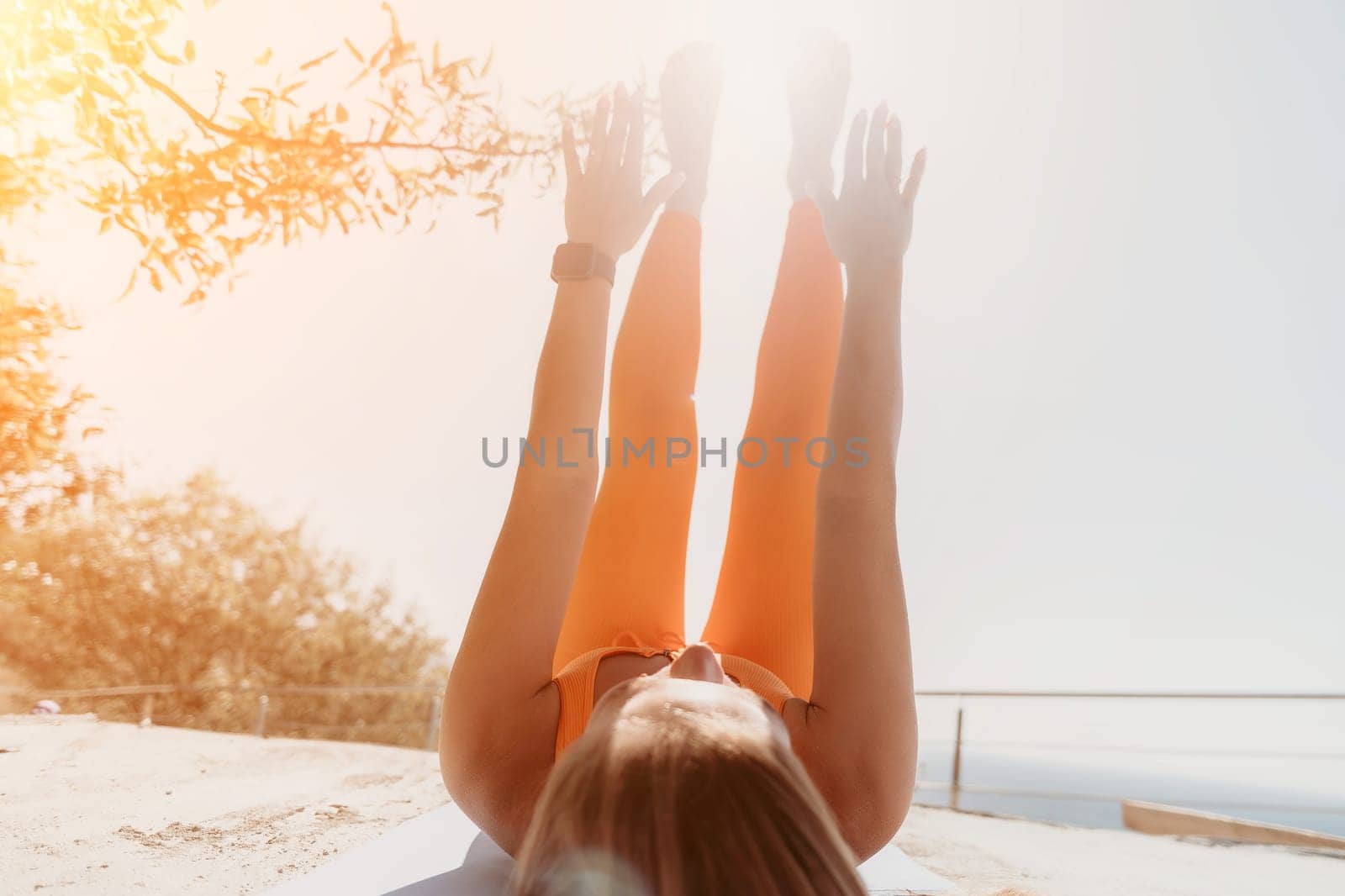 Fitness woman sea. Happy middle aged woman in orange sportswear exercises morning outdoors on yoga mat with laptop in park over ocean beach. Female fitness pilates yoga routine. Healthy lifestyle. by panophotograph