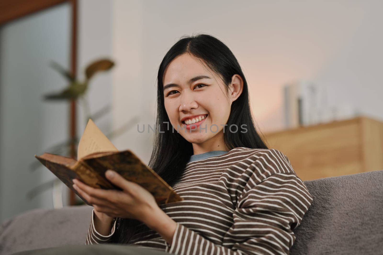 Young smiling woman wearing casual clothes reading book on grey sofa at home by prathanchorruangsak