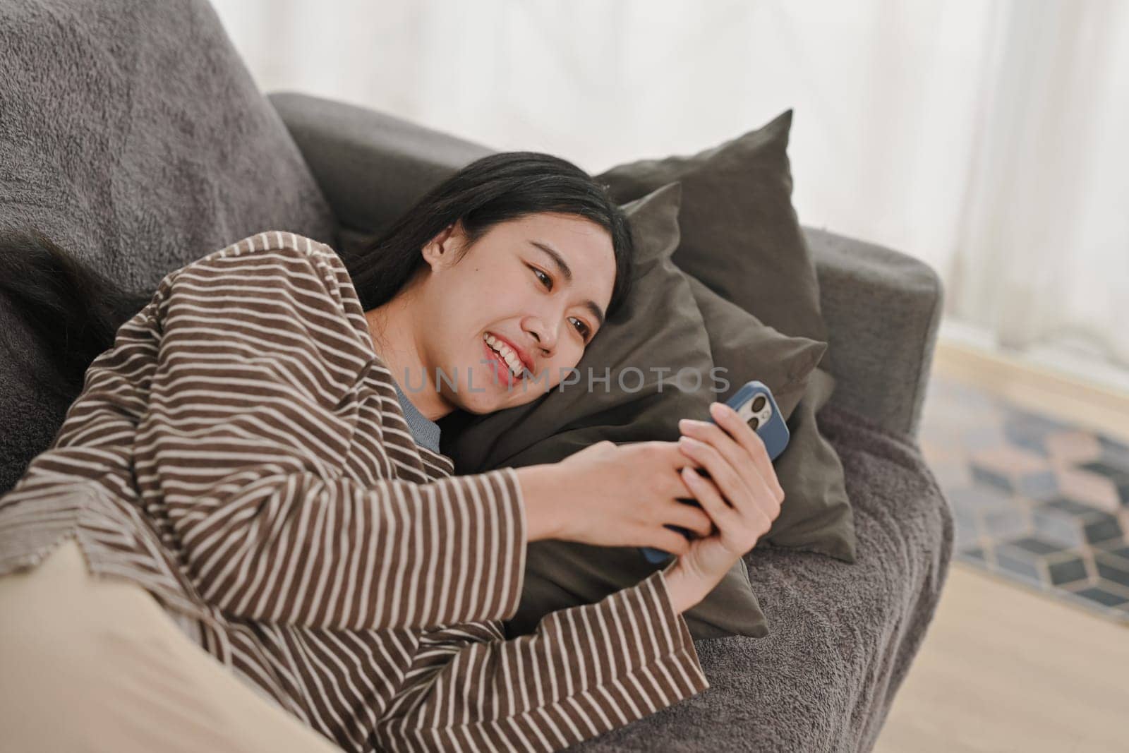 Smiling young woman in casual clothes lying on couch and using mobile phone.