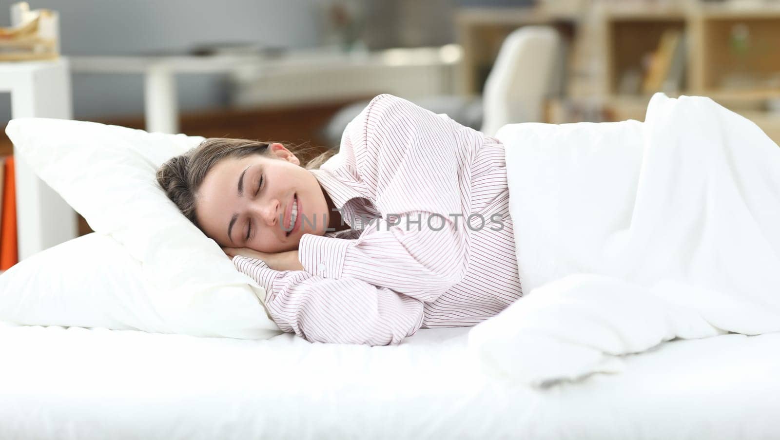 Happy girl in pajamas lies in bed closing her eyes. Woman is disturbed by insomnia, normalization sleep. Girl sleeps at home under white blanket. Healthy sleep during self-isolation