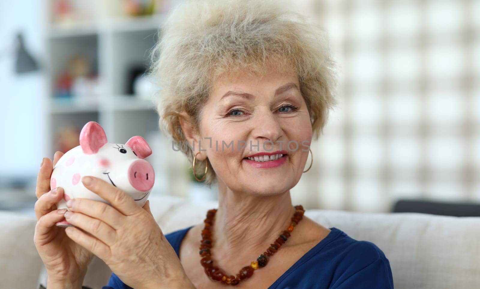 Happy elderly woman in apartment shows piggy bank. Accumulation pension capital. Elderly woman happily saving money in piggy bank. Older generation is used to saving and saving money