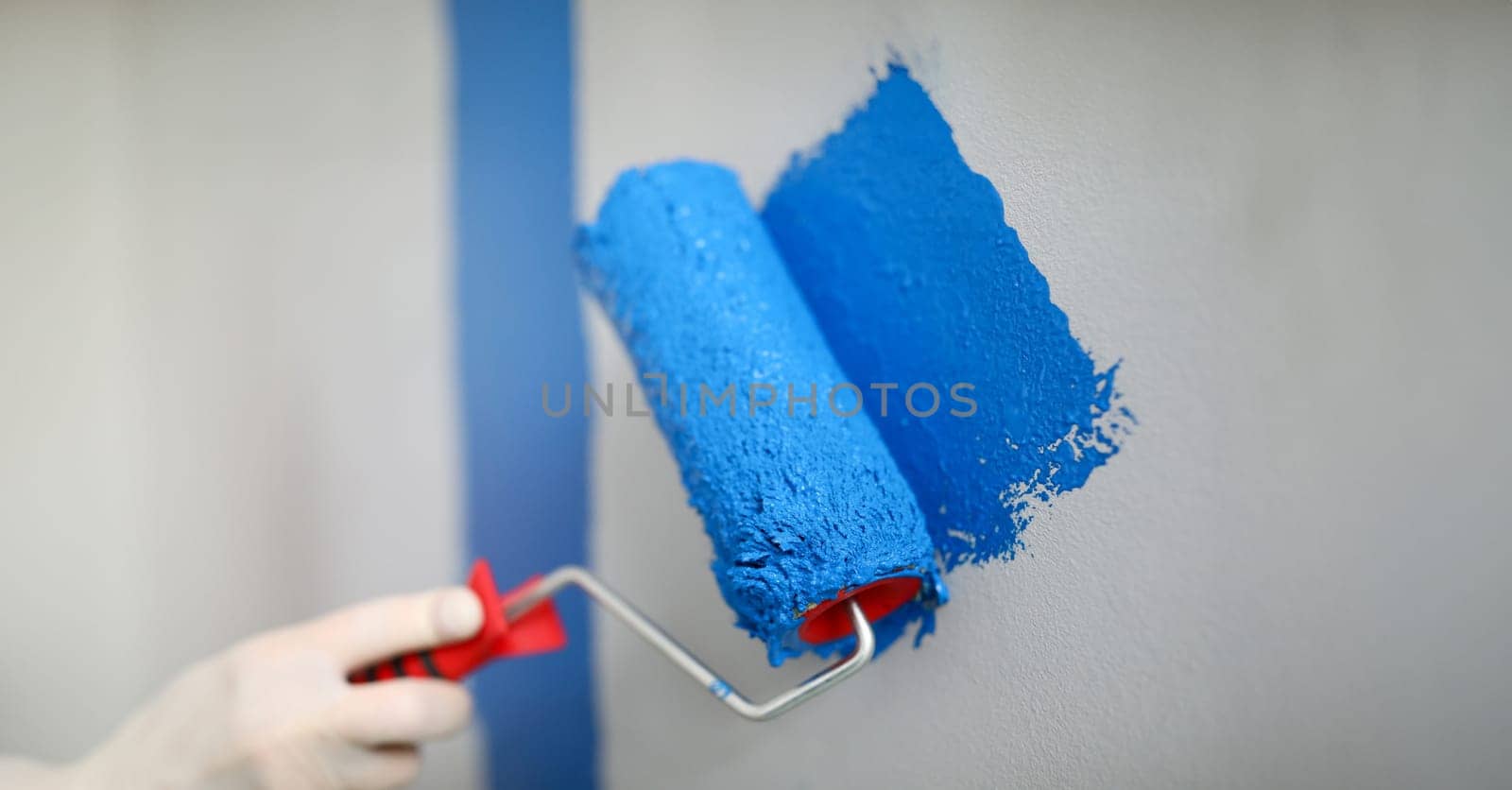 Hand holds roller and wire on wall with blue paint. Low roller performance. Convenient and versatile tool for painting walls. Tools with which best lay paint on surface. DIY repair in apartment