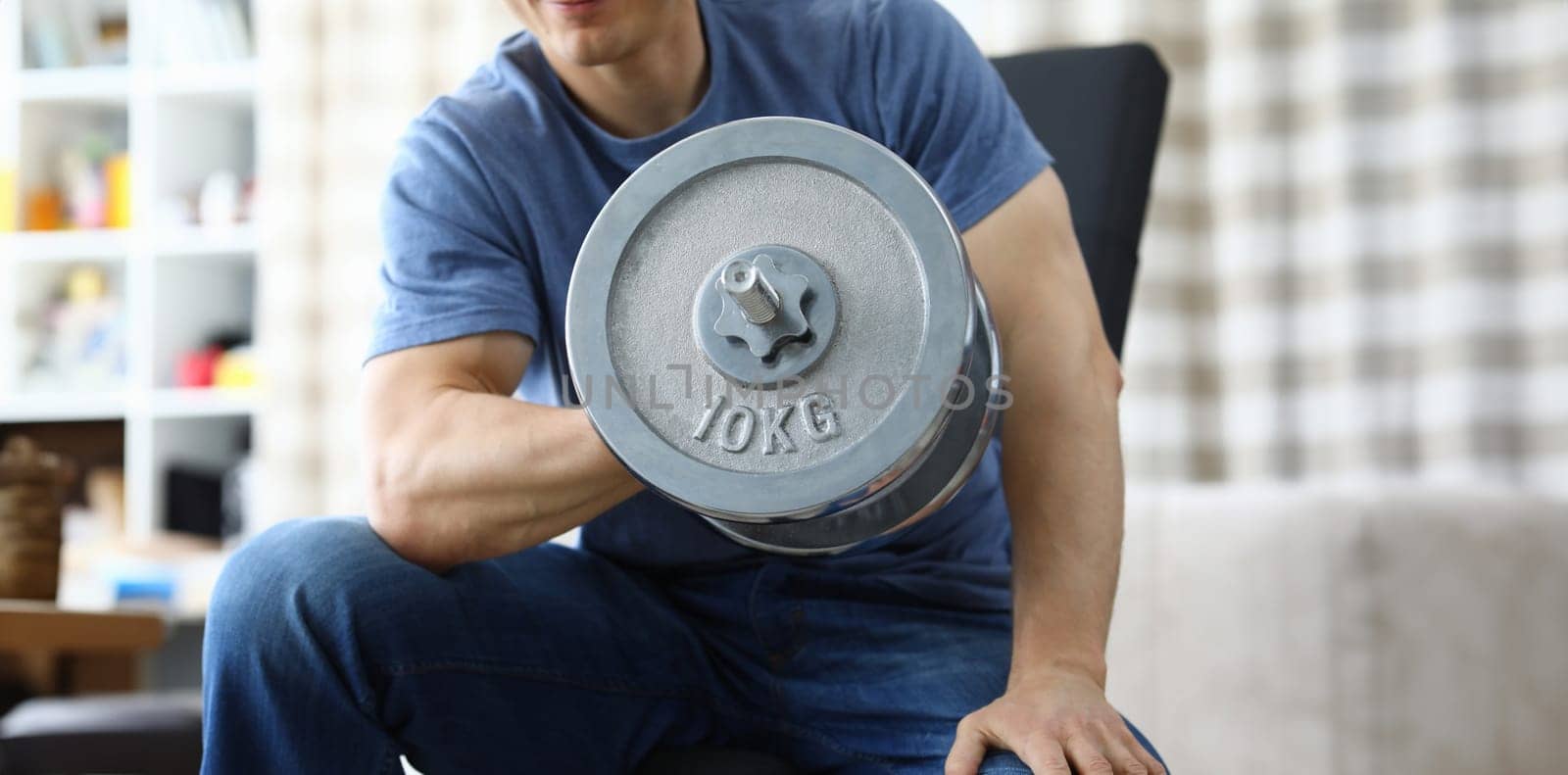 Man lifts heavy dumbbell while sitting on sofa by kuprevich