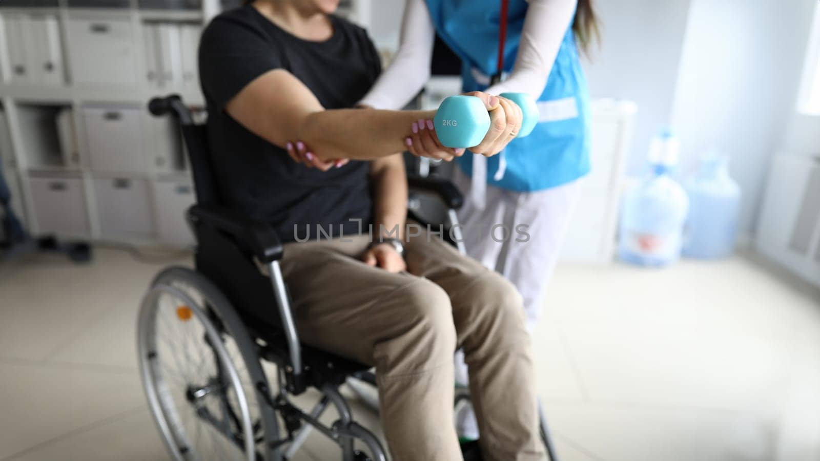Man on wheelchair works out his hand with dumbbell. Simple exercises for disabled. Doctor helps restore health. Exercise is suitable for people with disabilities who have power in their upper body