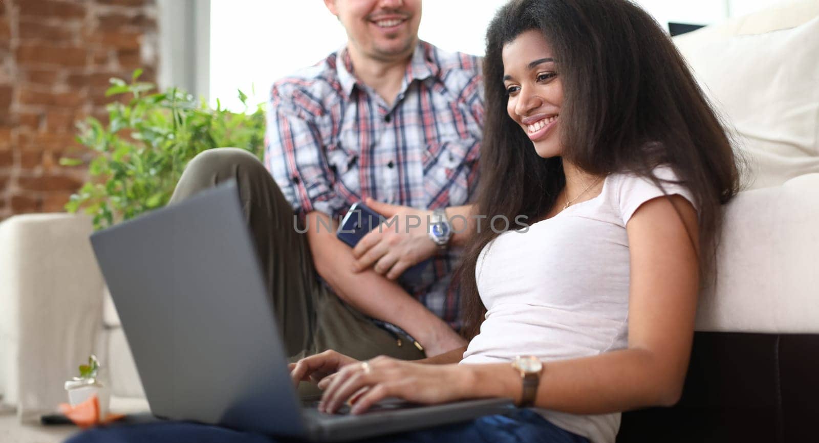 Smiling girl sitting on floor with laptop near guy. Guy and girl are looking for information on Internet and laughing. Husband looks at his wife with love. Dark-skinned girl working on laptop