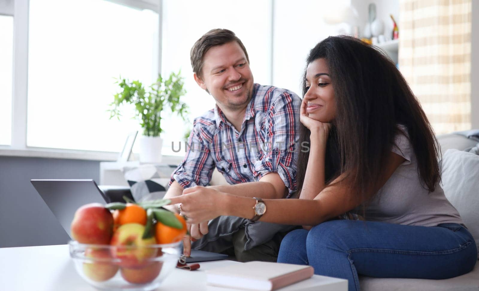 Happy couple talking at home at table on couch. Husband and wife at home looking at each other. Beautiful smiling girl touches fruit on table. Joyful discussion family issues at home