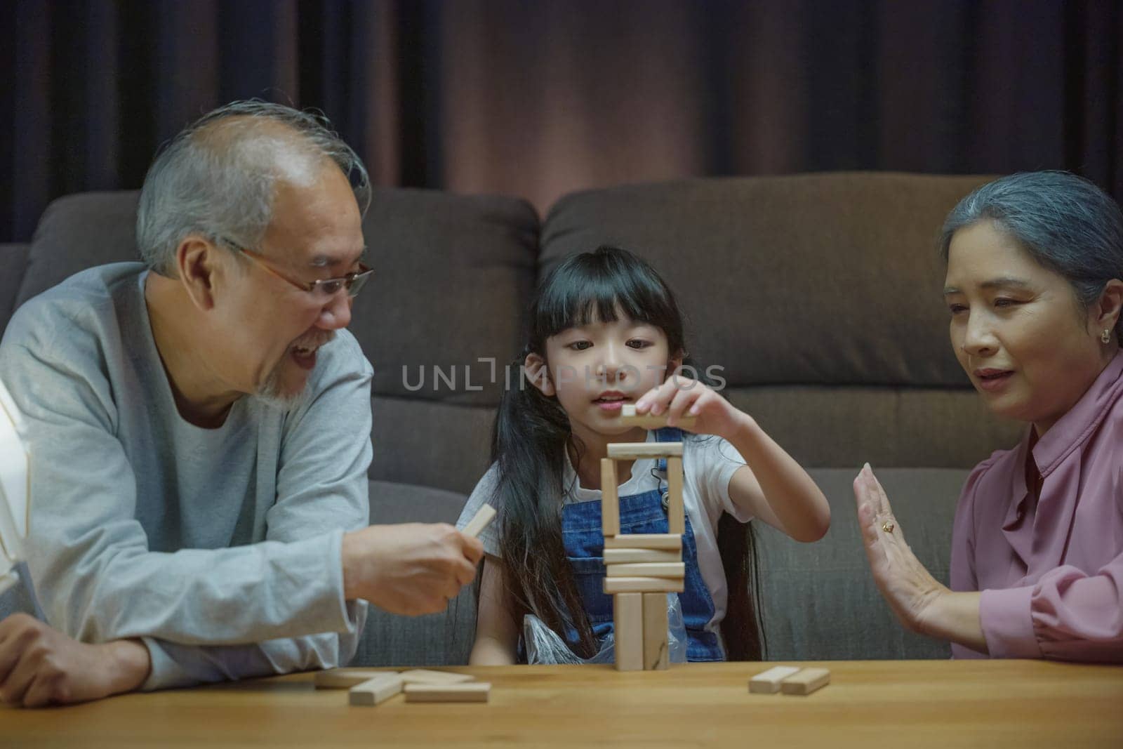 Happy grandparents Asian family enjoy playing toy block with little daughter together in home living room at night time, Smiling parent having fun play build constructor tower of wooden blocks