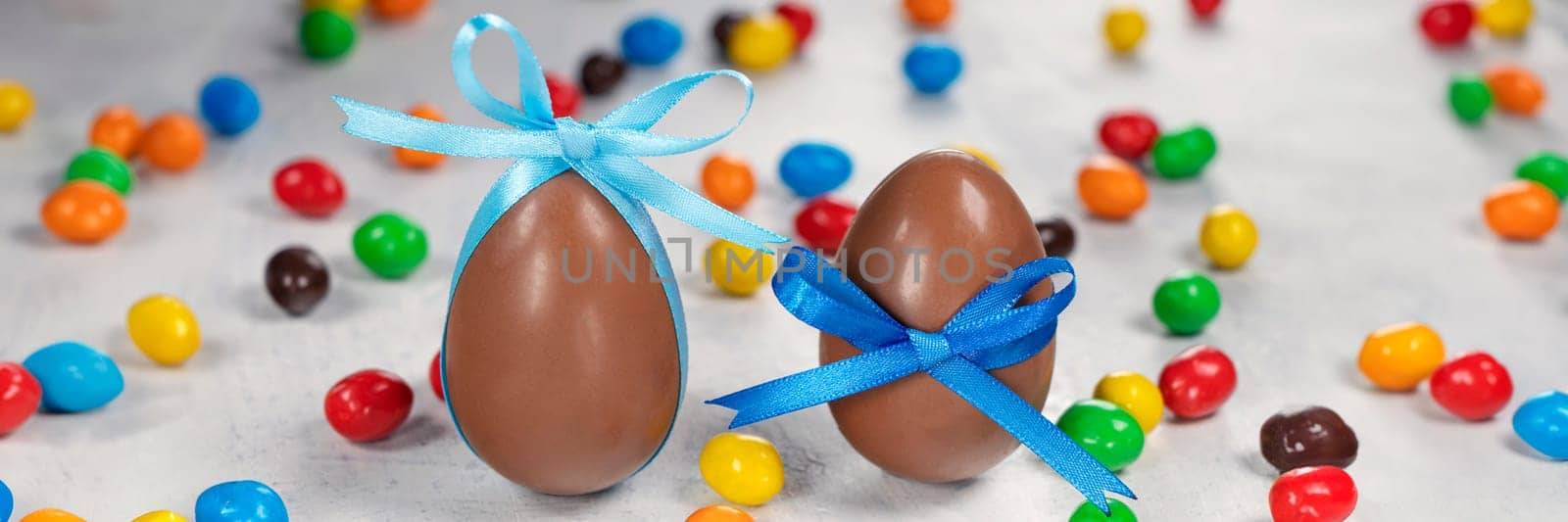 banner of Two big chocolate eggs with a blue bow on a white concrete background with colorful little eggs. by Leoschka