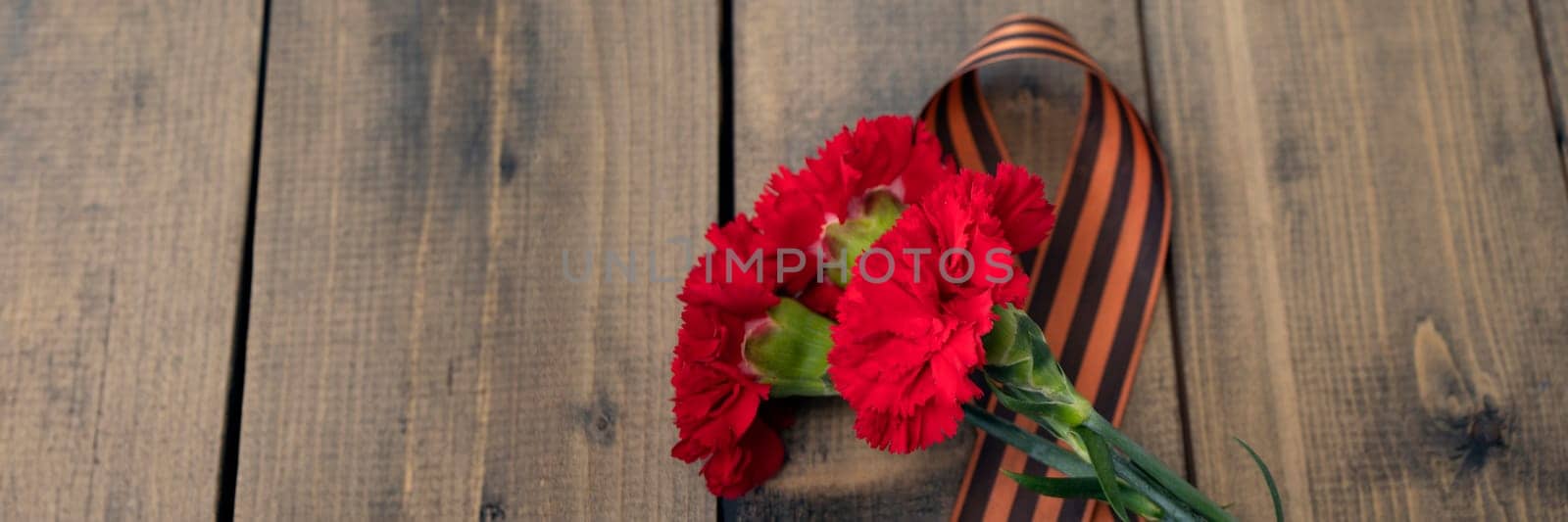banner of three red carnations with a St. George ribbon on a wooden background. Georgievsk ribbon - Russian symbol of victory and three red carnations. Holiday card for the holiday of May 9. by Leoschka
