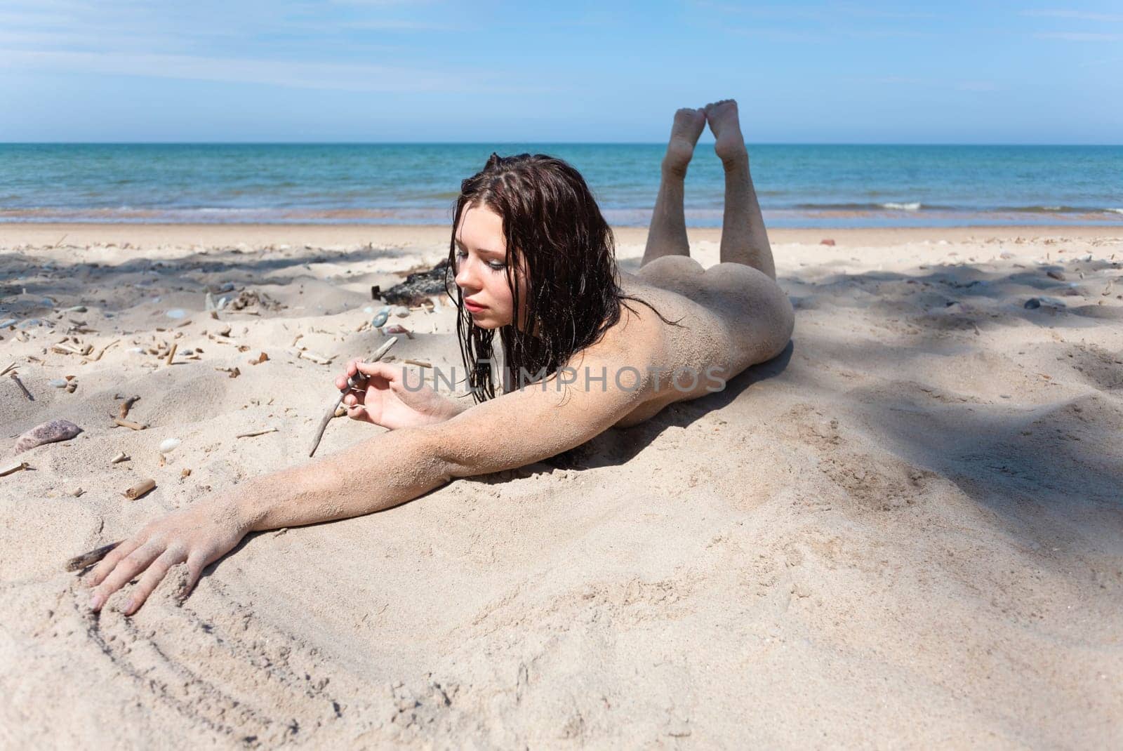 Youth, beauty, healthy lifestyle and nudity. Young fully naked woman with body covered with sand on the sea coast on a sunny day.