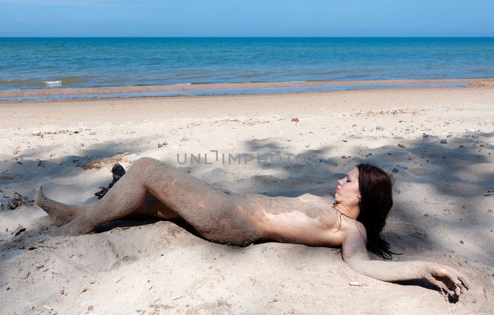 Youth, beauty, healthy lifestyle and nudity. Young fully naked woman with body covered with sand on the sea coast on a sunny day.