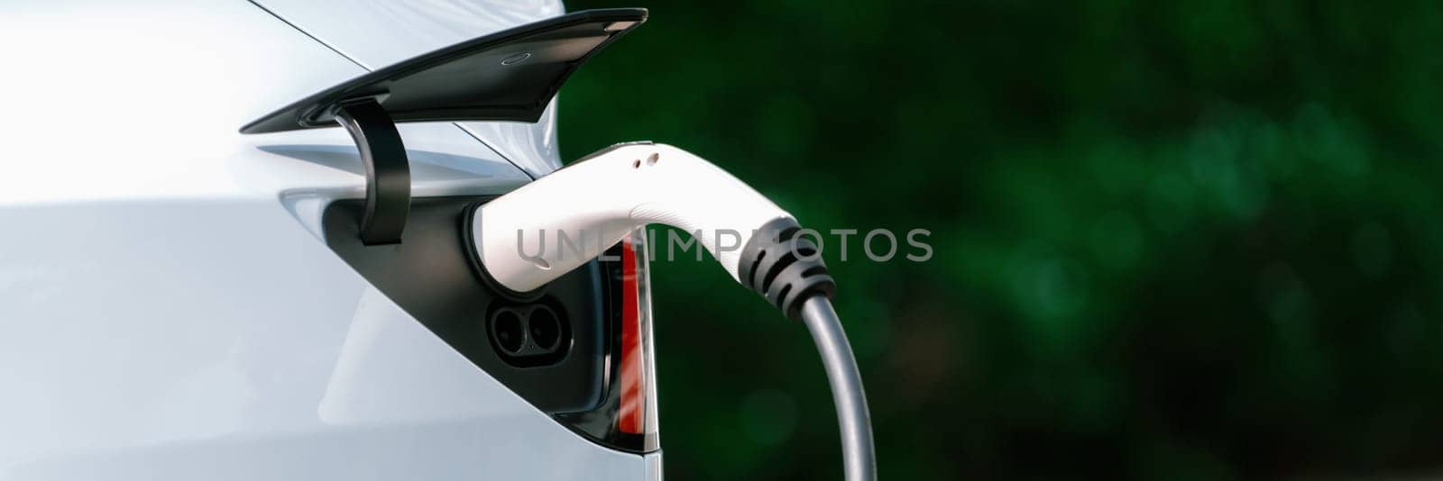 Closeup EV charger plug handle attached to electric vehicle port, recharging battery from charging station. Modern designed EV car and clean energy sustainability for better future.Panorama Synchronos