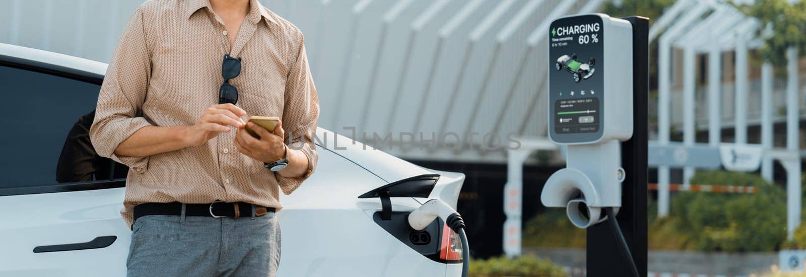 Young man use smartphone to pay for electricity at public EV car charging station at city commercial mall parking lot. Modern environmental and sustainable urban lifestyle with EV vehicle. Expedient