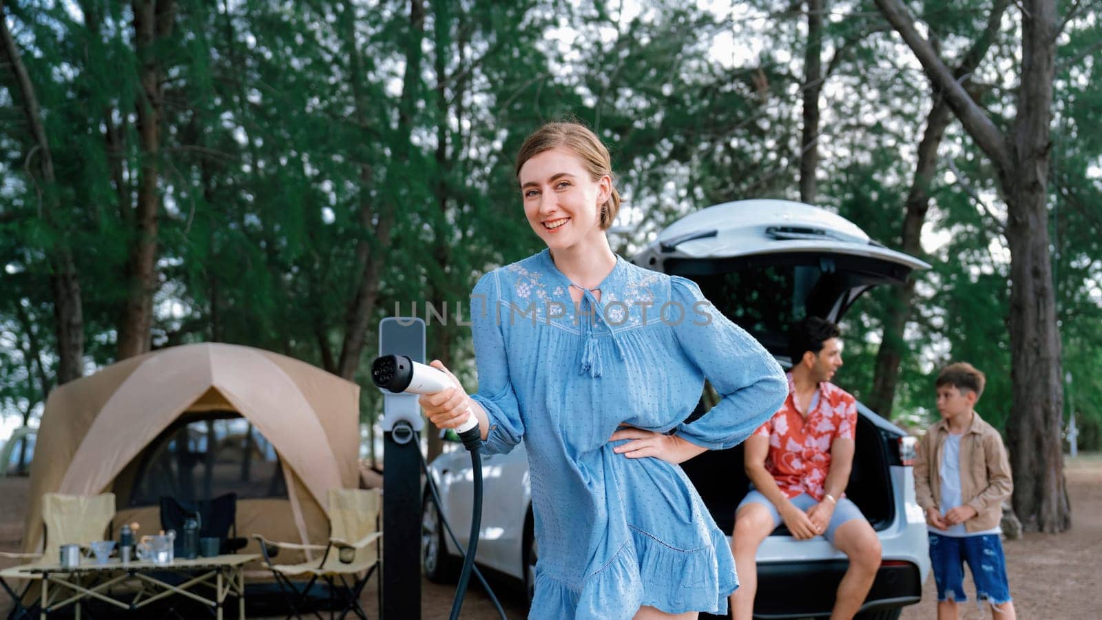 Outdoor adventure and family vacation camping at sea travel by eco friendly car. Cheerful woman or mother holding, pointing EV charger point with playful and happiness posture in campsite. Perpetual