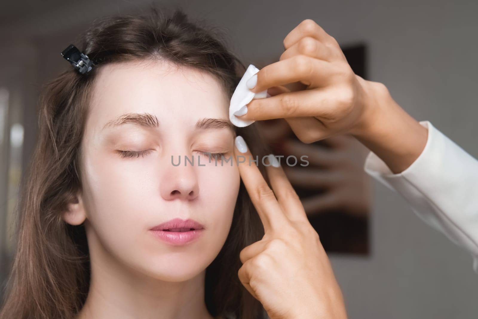 In a beauty salon, a master cosmetologist applies makeup and wipes eyebrows with a cotton pad. Close-up of the model's face during the procedure.
