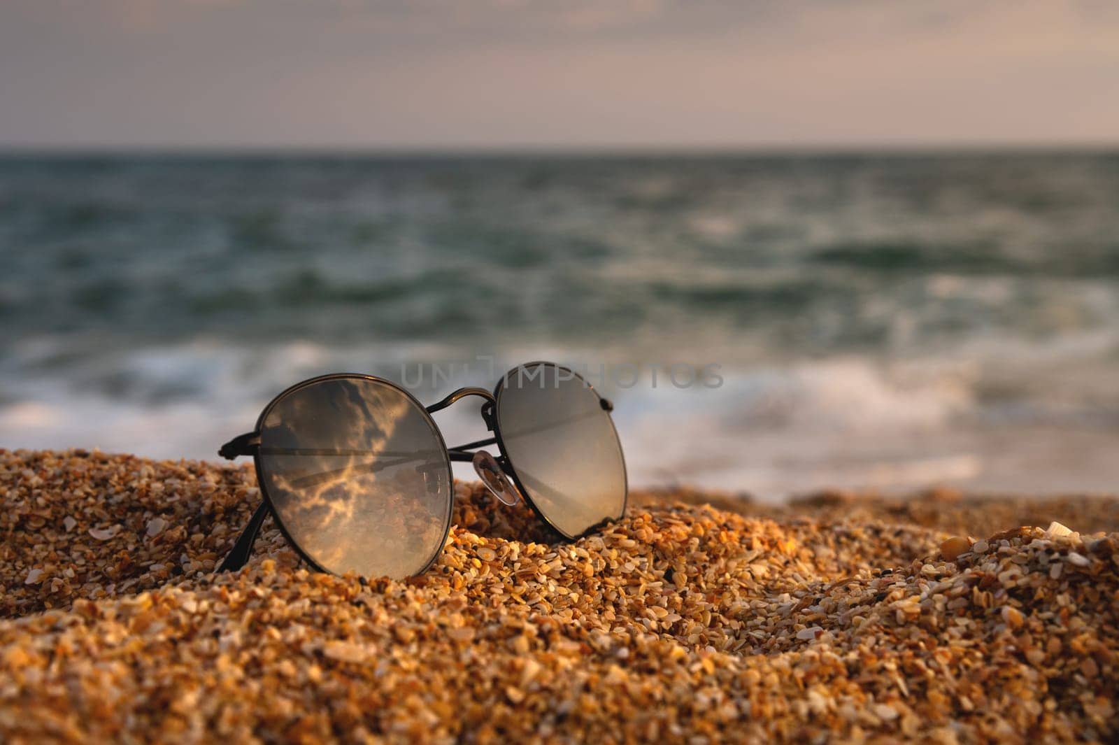 Side view of sunglasses on the beach near the turquoise sea with sand. Summer, beach holiday, stylish sunglasses, no people.