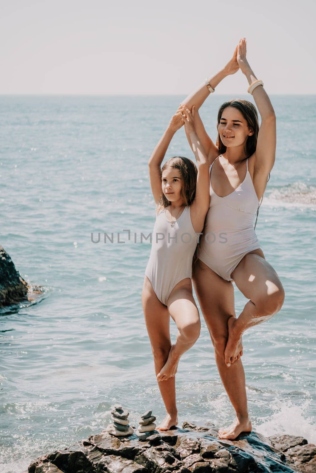 Woman and her daughter practicing balancing yoga pose on one leg up together on rock in the sea. Silhouette mother and daughter doing yoga at beach by panophotograph