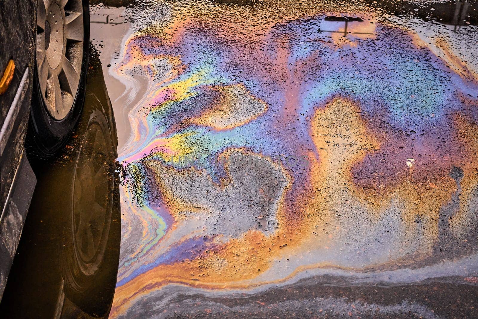 Spill of gasoline and oil on wet asphalt in a parking lot with the reflection of a car wheel in a puddle