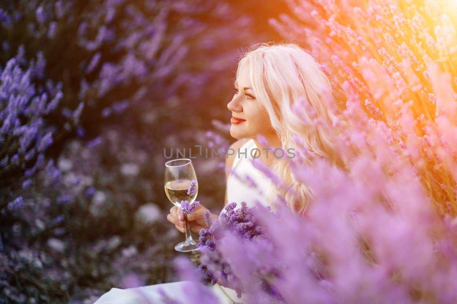 Blonde lavender field holds a glass of white wine in her hands. Happy woman in white dress enjoys lavender field picnic holding a large bouquet of lavender in her hands . Illustrating woman's picnic in a lavender field